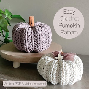 May include: Two crochet pumpkins, one lavender and one cream, sit on a wooden surface. The lavender pumpkin has a cinnamon stick stem. The cream pumpkin is decorated with a bow and leaf. The text "Easy Crochet Pumpkin Pattern" is visible.