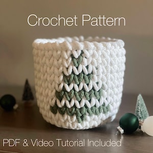 May include: A white crochet basket with a green Christmas tree design. The text "Crochet Pattern" is at the top, and "PDF & Video Tutorial Included" is at the bottom. The basket is surrounded by Christmas ornaments.