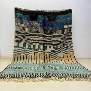 May include: A colorful, patterned rug with a mix of geometric designs. The rug features a palette of black, blue, teal, and beige. The design includes zigzags, stripes, and checkered patterns. The rug has a fringed edge.