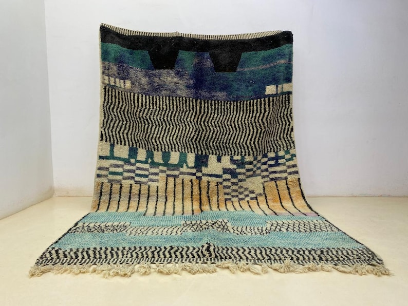 a large, colorful rug with a geometric pattern in shades of blue, green, and black. The rug is placed on a white floor against a white wall, creating a striking contrast.