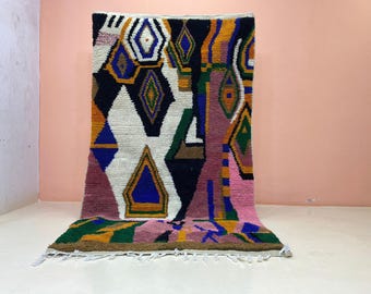 May include: A rectangular area rug featuring a bold, abstract geometric pattern in black, white, blue, green, orange, and pink. The design includes various diamond and triangular shapes throughout the surface, finished with white fringe at the bottom.