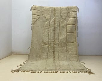 Puede incluir: Alfombra beige de estilo marroquí con un patrón geométrico. La alfombra presenta un diseño texturizado con secciones elevadas y flecos en el borde inferior. La alfombra es rectangular y parece estar hecha de un material suave y natural.