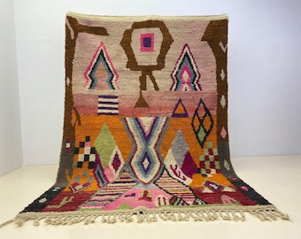 May include: A rectangular wool rug featuring a vibrant, multi-colored geometric pattern. The design includes shades of orange, pink, brown, purple, and green with various shapes like diamonds, triangles, and stripes. It has a tasseled fringe edge.