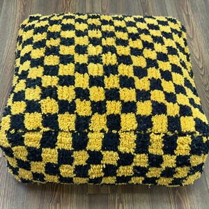 May include: Square pouffe with a black and gold chequered design. The textured fabric gives a plush look. This floor cushion adds a modern touch to any living space. Dimensions are approximately 50cm x 50cm.
