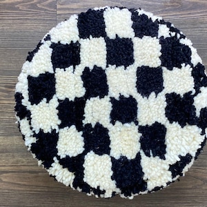 Pode incluir: Um pufe redondo com um padrão xadrez preto e branco. O pufe é feito de um material macio e texturizado, com um padrão de quadrados pretos e brancos alternados. O pufe é um item decorativo que pode ser usado como apoio para os pés ou assento extra.