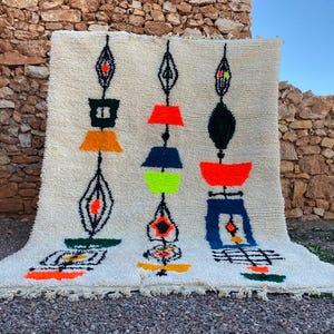 May include: Cream-coloured, hand-tufted rug with a geometric design. The rug features three vertical columns of abstract shapes in orange, black, blue, green, and red. The rug has a fringed edge and is displayed outdoors.