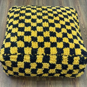 May include: A square pouffe featuring a yellow and black chequered design. The pouffe is made from a textured, plush fabric and is placed on a wooden floor. This is a bold, geometric piece for home decor, measuring approximately 60cm x 60cm.
