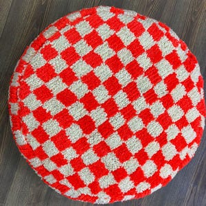 May include: Round, red and white checkered floor cushion. The cushion has a plush, textured surface with a repeating pattern of red and white squares. The cushion is circular and appears to be made of a soft, woven material.