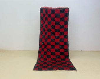 Op de afbeelding: Een rechthoekig, handgeweven tapijt met een opvallend rood en zwart dambordpatroon. Het tapijt is gemaakt van een dik, getextureerd materiaal en heeft aan de bovenkant een franje. Het tapijt wordt gepresenteerd tegen een neutrale achtergrond.