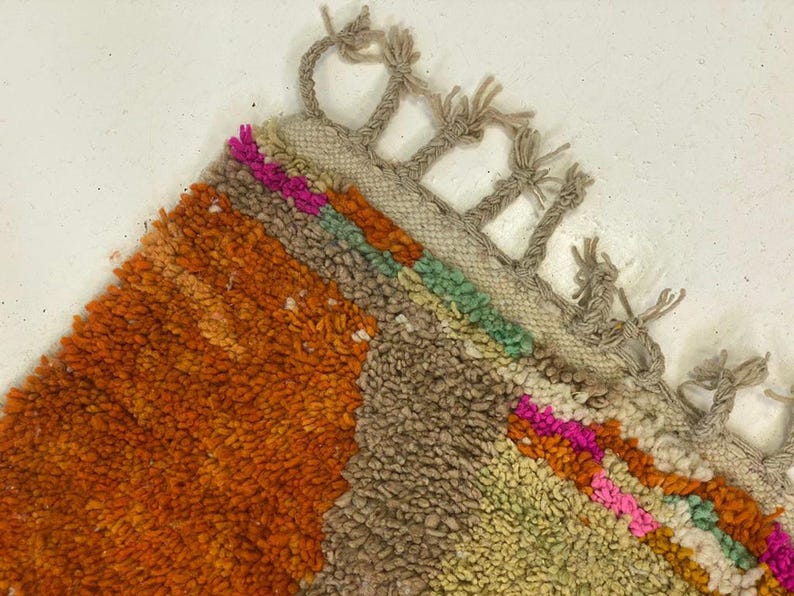 a vibrant, multicolored rug with a geometric pattern of orange, green, and pink hues. The rug has a fringe made of small, multicolored strings that add texture and visual interest to the overall design.