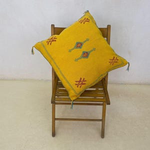 May include: A square, mustard-yellow pillow with embroidered geometric designs in teal, orange, and red. The pillow is placed on a wooden chair. The pillow has tassels on each corner. The background is a white wall and a light-colored floor.