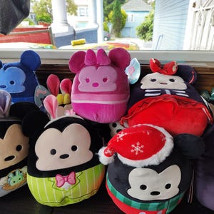 May include: A collection of plush toys featuring Mickey and Minnie Mouse characters in various designs. The toys include Easter bunny ears, a Santa hat, and other themed outfits. The plushies are in shades of black, blue, pink, red, and green.