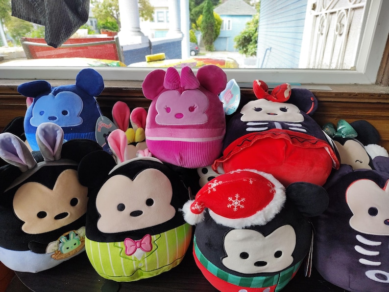 May include: A collection of plush toys featuring Mickey and Minnie Mouse characters in various designs. The toys include Easter bunny ears, a Santa hat, and other themed outfits. The plushies are in shades of black, blue, pink, red, and green.
