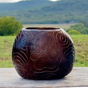 May include: A dark brown, round, carved wooden bowl with a bear paw print design. The bowl has a wide opening and is decorated with swirling patterns. The background shows a green field and a distant mountain range.