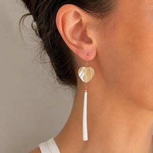 May include: A drop earring featuring a heart-shaped iridescent shell charm suspended from a rose gold-toned hook. Attached to the bottom of the heart is a long, slender, white cylindrical shell pendant that hangs vertically from a small metal bead.