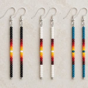 May include: Three pairs of long, linear drop earrings featuring vertical strands of small seed beads. The first pair is black, the second white, and the third teal, each accented with a central pattern of red, orange, yellow, and white beads.