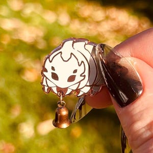 May include: A white and gold enamel pin featuring a stylized insect design with a small bell charm. The pin has a detailed design with a gold outline and a white interior. The bell charm is a small, bronze-colored bell.