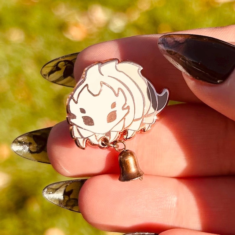 May include: A gold-colored enamel pin featuring a stylized, cream-colored creature with black and white stripes. The pin has a small, dangling bell. The pin is held between fingers against a blurred green background.