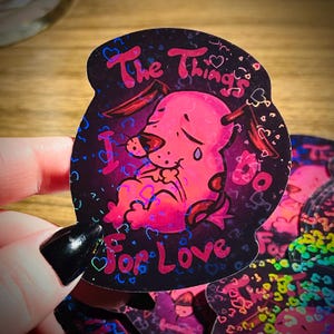 May include: A pink cartoon dog with a sad expression is featured on a black sticker with a holographic finish. The sticker says "The things I do for love" with hearts scattered around the dog.