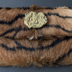 Könnte beinhalten: Rechteckige Clutch mit Tigerstreifenmuster in Braun und Schwarz. Sie hat einen goldfarbenen Metallverschluss mit detailliertem Drachendesign. Die Tasche ist aus einem weichen, strukturierten Material gefertigt.