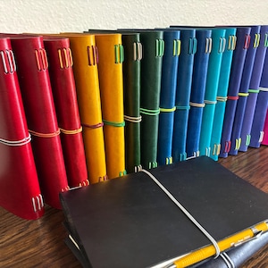 May include: A collection of colorful leather journals in various shades, including red, yellow, green, blue, and purple. Each journal is bound with a contrasting elastic band and white stitching. A black journal with a yellow pen lies in front of the standing journals.