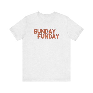 Pu&ograve; includere: T-shirt grigio chiaro con la scritta arancione "SUNDAY FUNDAY". Il testo &egrave; in un carattere giocoso e la maglietta sembra essere fatta di un materiale morbido e confortevole.