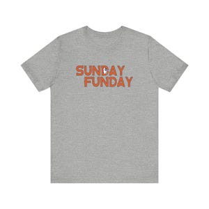 Pu&ograve; includere: T-shirt grigia con la scritta arancione "SUNDAY FUNDAY". Il testo &egrave; in un carattere giocoso, con una piccola grafica al centro della parola "FUNDAY".