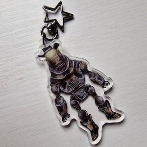 May include: A clear acrylic keyring with a robot cowboy design. The robot is depicted in brown and grey, wearing a hat and boots. The keyring has a black star-shaped clasp.