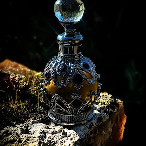 Puede incluir: Un frasco de perfume ornamentado con un diseño de filigrana plateada y un tapón de cristal facetado. El interior del frasco es amarillo dorado y se encuentra sobre un fondo oscuro, resaltando los detalles.