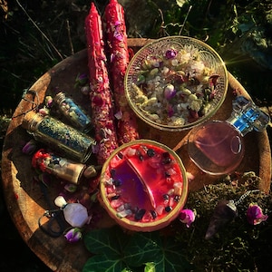 Peut inclure: Un bol en bois rempli de divers objets pour un rituel ou un sort. Il y a deux bougies rouges, un petit bol de sel rose et blanc, une bouteille en verre transparente avec un liquide rose, plusieurs petites bouteilles en verre avec des cristaux colorés, une bougie rose avec des cristaux incrustés et un pendule.