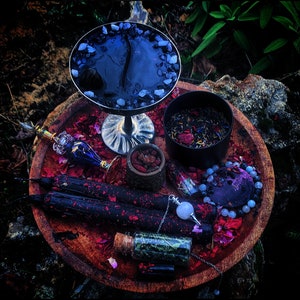 Peut inclure: Un plateau en bois contenant divers objets pour un rituel spirituel, notamment un bol noir avec des cristaux, un petit bol d'herbes, une bouteille d'huile, un pendule et deux bougies noires.