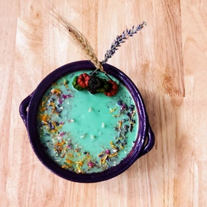 Op de afbeelding: Een turquoise kaars in een dieppaarse keramische kom. De kaars is versierd met gedroogde bloemen en kruiden. Een takje lavendel en tarwe zijn bovenop de kaars geplaatst. De kom heeft twee handvatten.