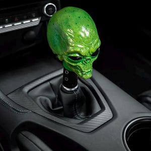 May include: A green alien head shift knob installed in a car. The shift knob is a detailed sculpture with black eyes and a textured surface. The interior of the car is black with a carbon fiber trim.