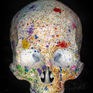 Puede incluir: Una calavera decorativa con base blanca, cubierta de salpicaduras de pintura coloridas. La pintura incluye rojo, azul, amarillo, morado y verde. La calavera tiene cuencas oculares grandes y una nariz y mand&iacute;bula detalladas. El fondo es negro.