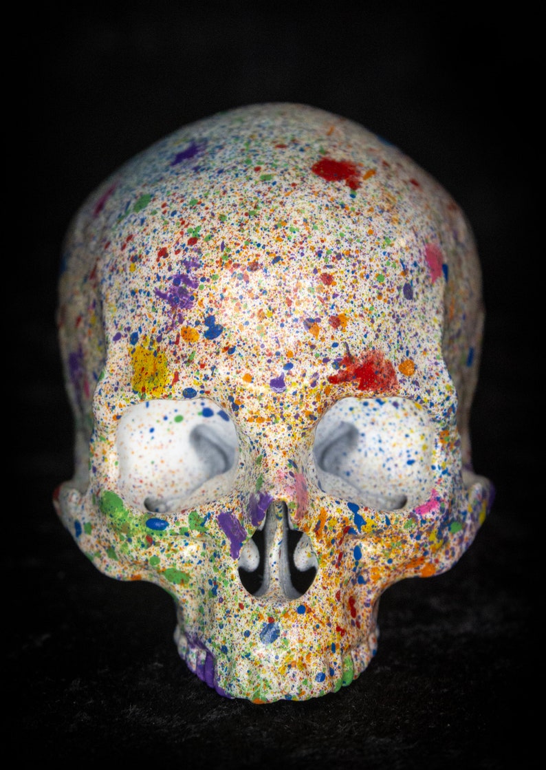 Puede incluir: Una calavera decorativa con base blanca, cubierta de salpicaduras de pintura coloridas. La pintura incluye rojo, azul, amarillo, morado y verde. La calavera tiene cuencas oculares grandes y una nariz y mand&iacute;bula detalladas. El fondo es negro.