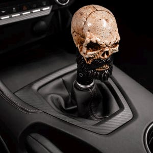 May include: A skull-shaped gear shift knob with a black beard and mustache. The skull is tan with dark accents, and the shift knob is installed in a black car interior. The overall theme is automotive accessories.