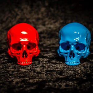 Puede incluir: Dos figuras de calaveras brillantes, una roja brillante y otra azul vibrante, se exhiben sobre un fondo oscuro y texturizado. Las calaveras son id&eacute;nticas en forma, con cuencas de los ojos y cavidades nasales visibles, mostrando un contraste llamativo.
