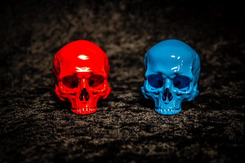 Puede incluir: Dos figuras de calaveras brillantes, una roja brillante y otra azul vibrante, se exhiben sobre un fondo oscuro y texturizado. Las calaveras son id&eacute;nticas en forma, con cuencas de los ojos y cavidades nasales visibles, mostrando un contraste llamativo.