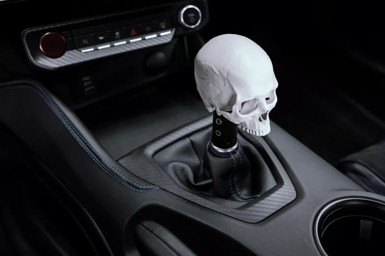 Puede incluir: Un pomo de palanca de cambios con forma de calavera blanca instalado en un coche. La calavera tiene cuencas de los ojos, una cavidad nasal y dientes detallados. El interior del coche es negro con costuras azules.