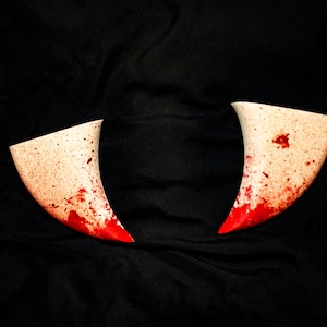 May include: Two white horns with red splatters of paint on a black background.