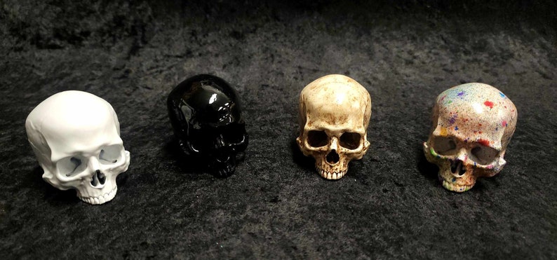 Puede incluir: Cuatro figuras decorativas de calaveras en fila. Una es blanca, otra negra brillante, otra color hueso y la &uacute;ltima salpicada de pintura colorida. Las calaveras son un elemento decorativo macabro.