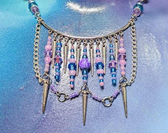 May include: A multi-strand necklace featuring a silver-toned curved bar with dangling silver spikes, blue and purple glass beads, and small light pink spheres. The design incorporates silver chains connecting the beaded segments in a layered arrangement.