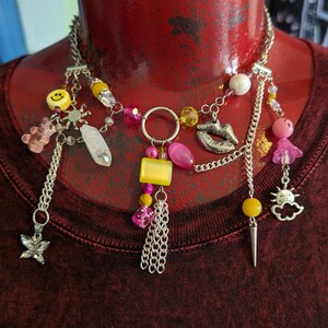 May include: A silver chain necklace with various charms, including a smiley face, a butterfly, a crystal, and pink and yellow beads. The necklace is displayed on a red mannequin head, with a maroon shirt in the background.