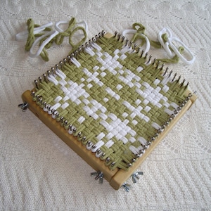 May include: A square weaving loom with a woven green and white pattern. The loom is made of wood and has metal pins around the edges. Several strands of green and white yarn are next to the loom.