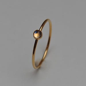 Op de afbeelding: Een fijne gouden ring met een kleine, iriserende edelsteen. De ring heeft een dunne band en een minimalistisch ontwerp. De edelsteen heeft een mix van kleuren, waaronder blauw, oranje en geel. De ring staat tegen een neutrale achtergrond.