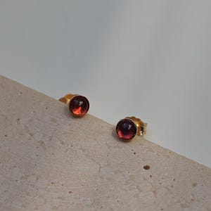 Pode incluir: Um par de brincos de ouro com tachas, cada um com uma pequena gema redonda, vermelho-alaranjada. Os brincos estão colocados sobre uma superfície clara e texturizada, contra um fundo suave e neutro. As pedras preciosas têm um ligeiro gradiente de cor.