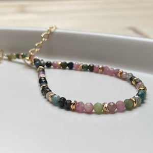 Puede incluir: Un brazalete de piedras preciosas multicolores con cuentas rosas, verdes y azules. El brazalete tiene un cierre dorado y est&aacute; enhebrado en una cadena dorada.