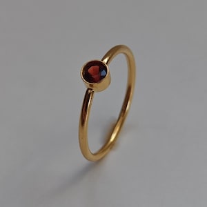 Könnte beinhalten: Ein dünner Goldring mit einem einzelnen, kleinen, runden, tiefroten Granat-Edelstein in einer einfachen Zargenfassung. Das Metall hat eine glatte, polierte Oberfläche und der Ring ist schräg positioniert, um den runden Stein zu zeigen.