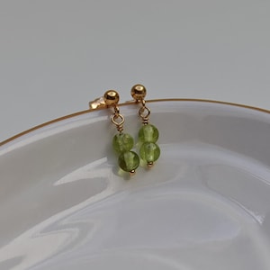 May include: A pair of gold-colored earrings with a post and a dangling design. Each earring features two light green, round beads. The earrings are displayed on a white, oval-shaped dish with a gold rim.