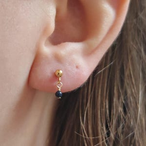Puede incluir: Un pendiente de botón dorado con una pequeña bola de oro pulido en el lóbulo, conectada por un hilo fino a una diminuta cuenta esférica de color azul oscuro que cuelga verticalmente bajo el botón, creando un diseño colgante sencillo.